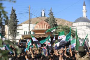 Syrians protest against sectarianism in 2024 with green Syrian flags being waved above a crowd of people.