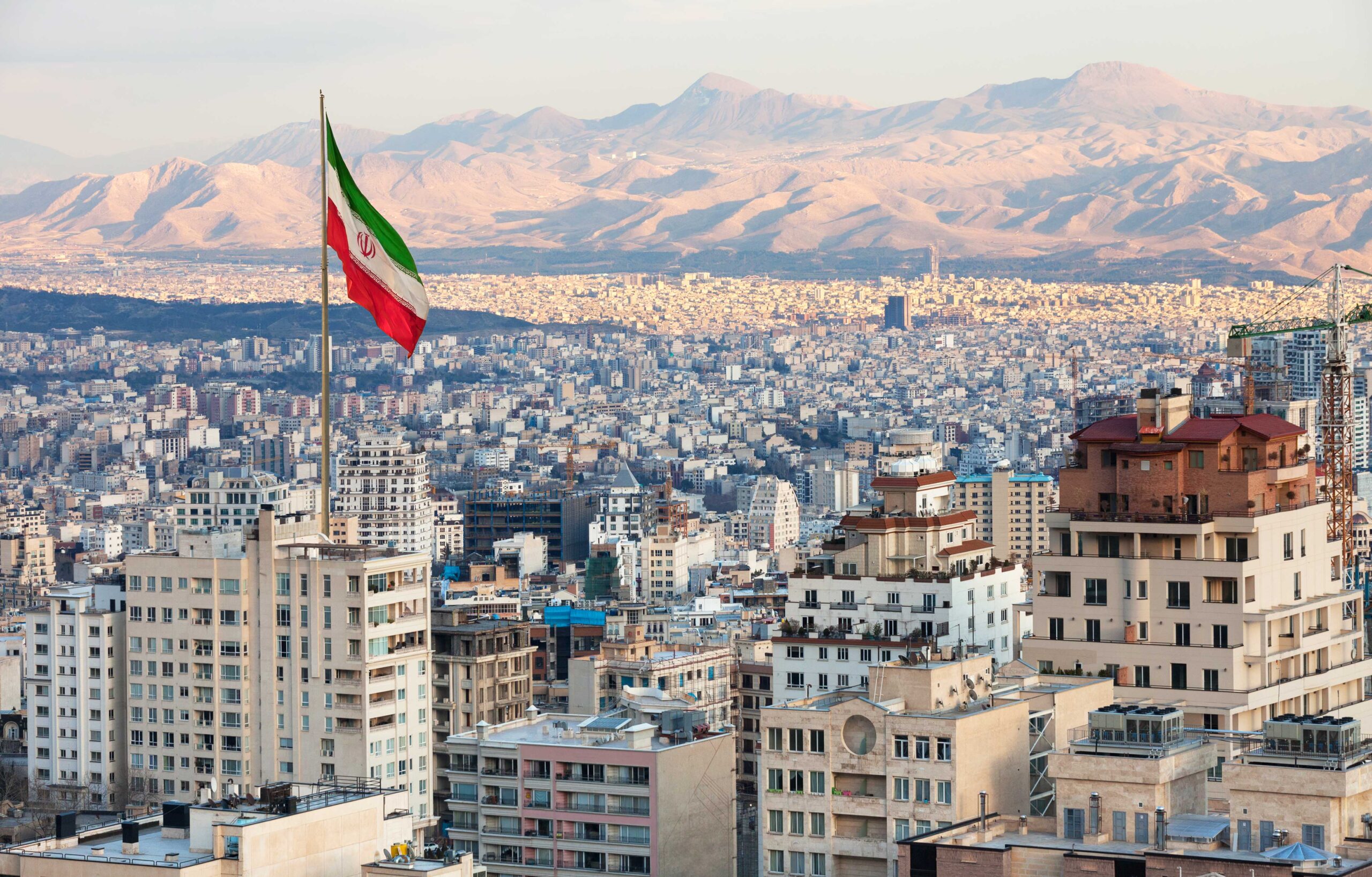 Waving Iran flag above skyline of Tehran at sunset.