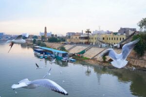 Baghdad, Iraq – December 1, 2025: A view of the Al-Qushla Clock Tower and boats along the Tigris River, framed through the railings of Al-Shaheed Bridge, with seagulls flying over the water.