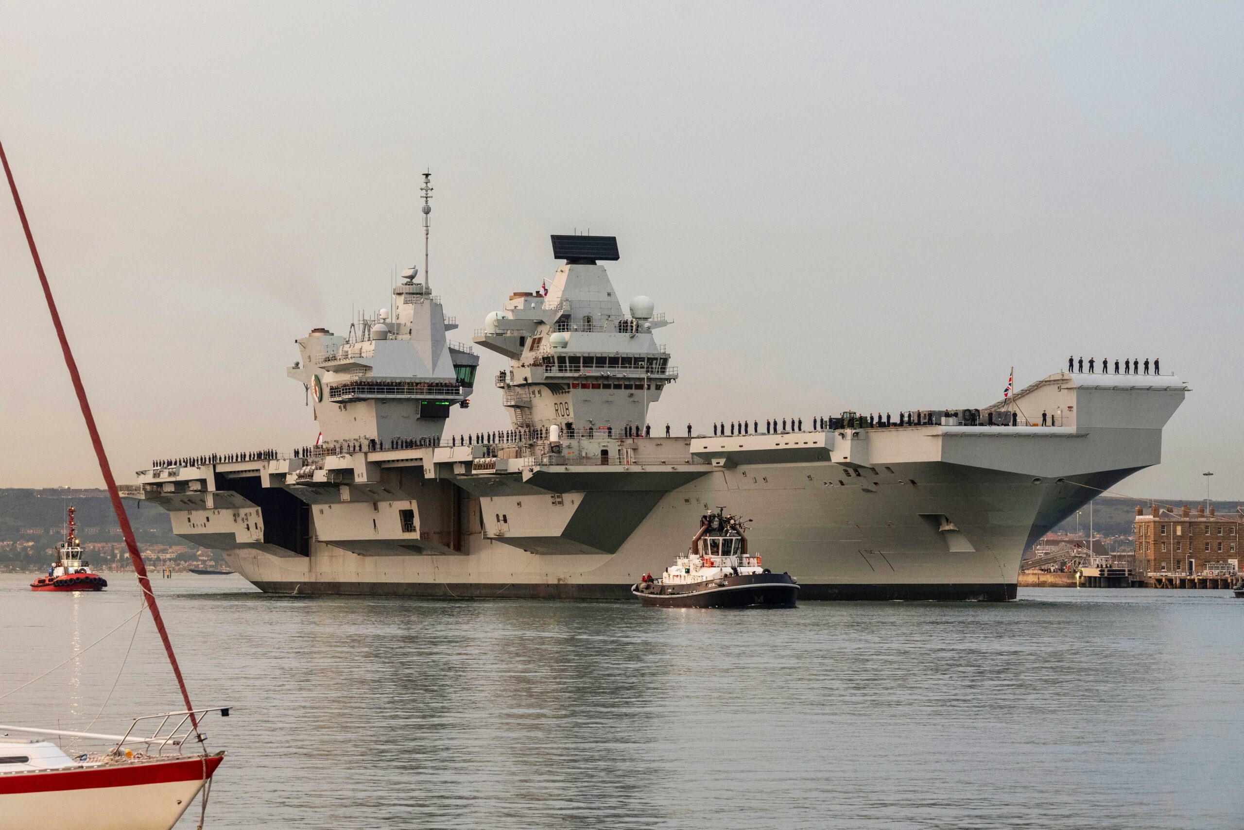 Portsmouth, England, UK. 8 September 2023. HMS Queen Elizabeth underway leaving Portsmouth dockyard at dusk with an escort of tugs and security vessels. Naval personnel line the decks.