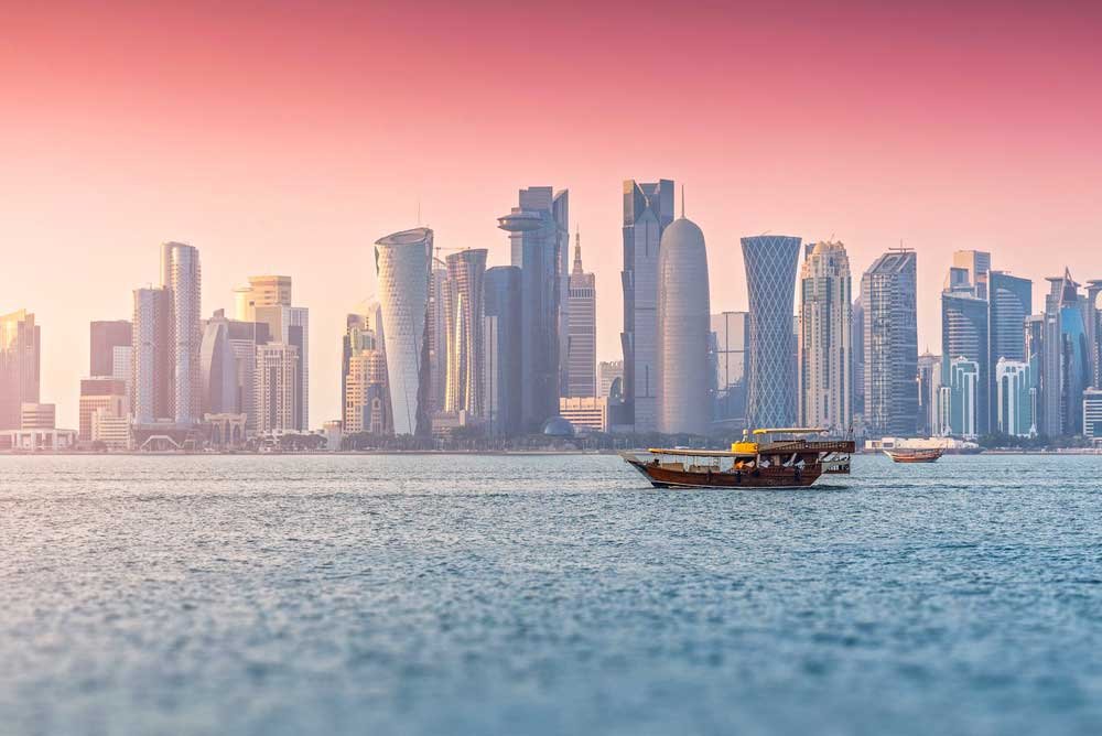 Doha city skyline featuring West Bay skyscrapers along the Persian Gulf during a beautiful pastel sunset, with dhow boats sailing